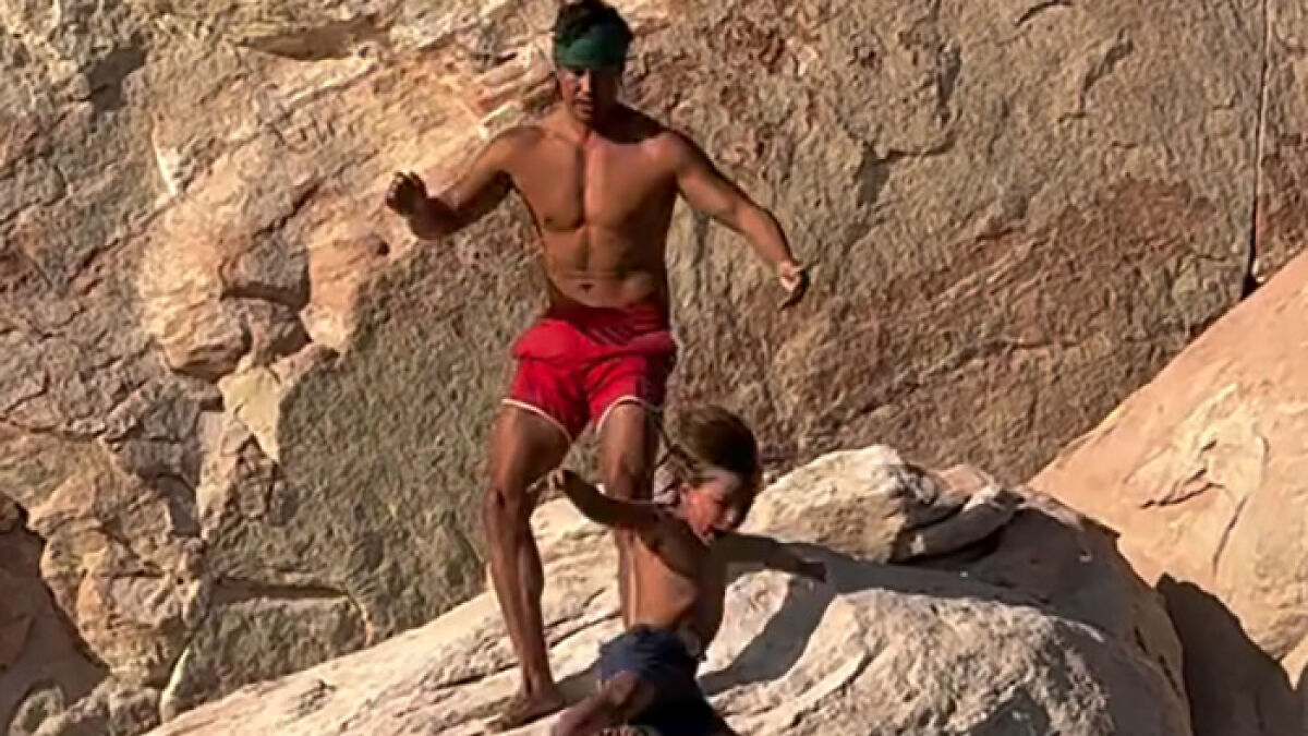 Man in red shorts throwing 7-year-old boy off cliff to teach him to face his fears near rocky terrain.