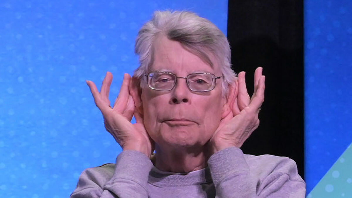 Stephen King wearing glasses and gray sweatshirt, making a gesture with hands near ears, related to Epstein list conspiracy theories.