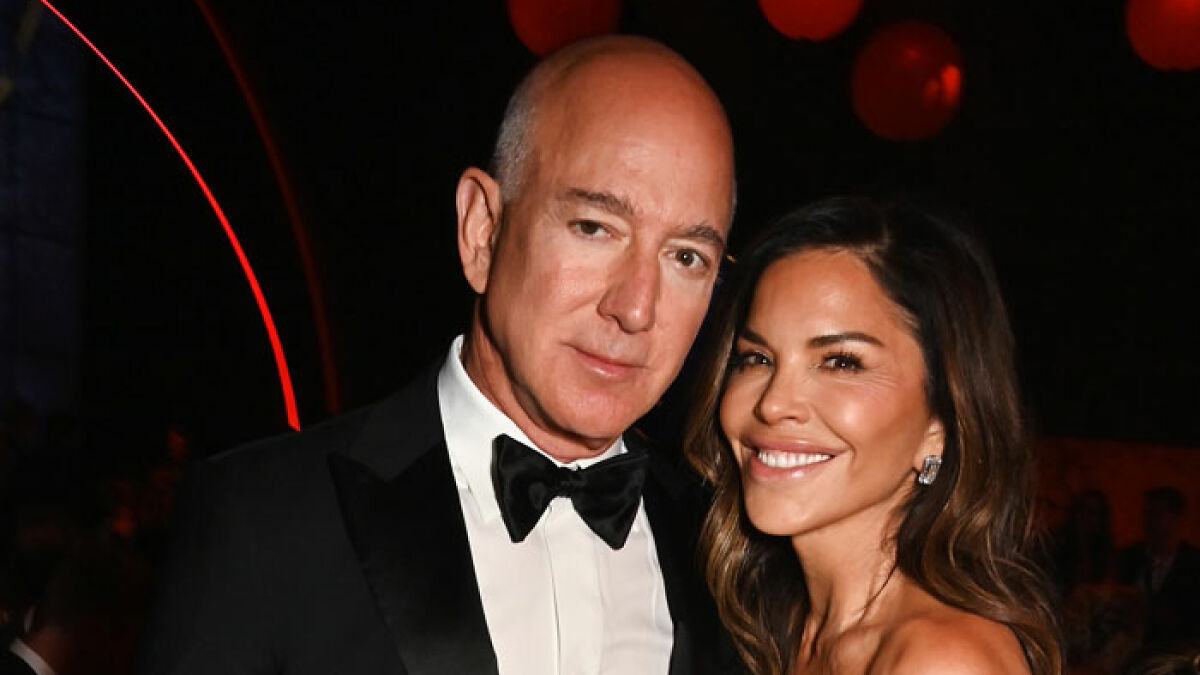 Jeff Bezos with new hair posing alongside smiling Lauren Su00e1nchez at a formal event in a dark background.