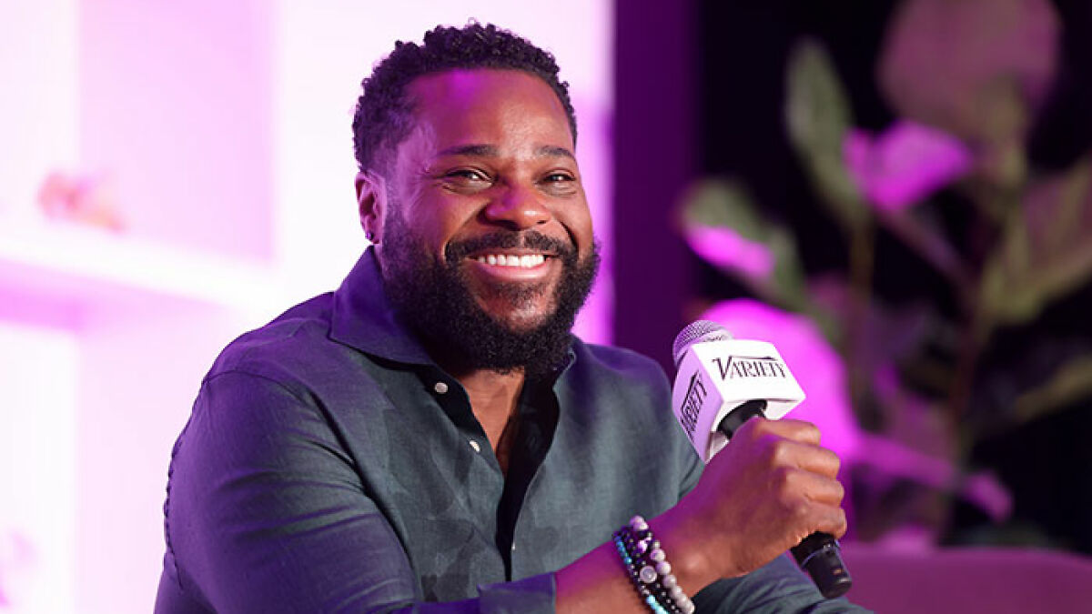 Malcolm-Jamal Warner smiling while holding a microphone during one of his final public appearances with purple lighting.
