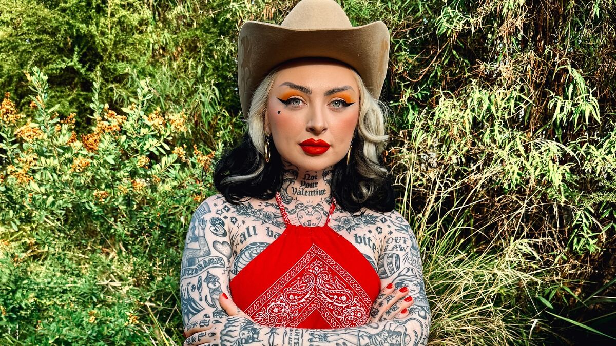 Woman with 250 tattoos as body suit transformation wearing a cowboy hat and red top standing outdoors.