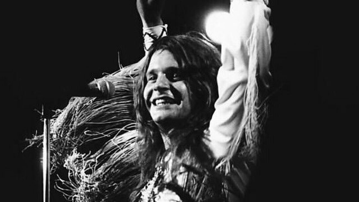 Black and white photo of Ozzy Osbourne performing on stage, smiling and raising his arm during a rock tribute.