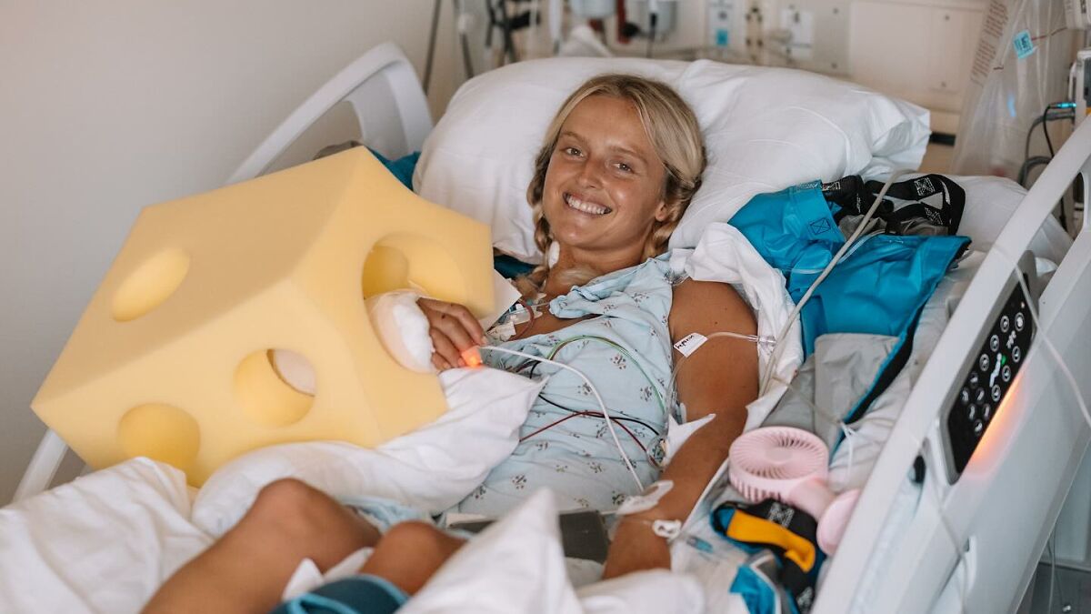 Surf instructor in hospital bed smiling with medical equipment and arm covered by a large foam block after injury.