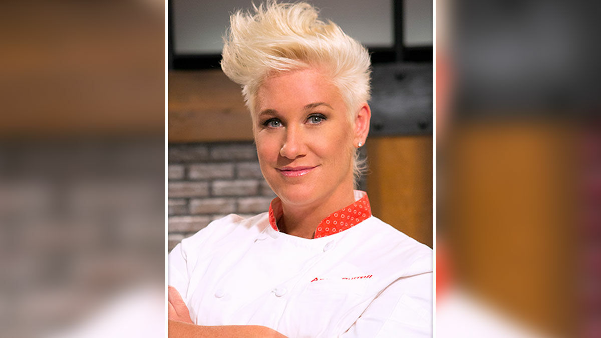Master Chef Anne Burrell in a white chef coat posing confidently indoors with styled short blonde hair and a slight smile.