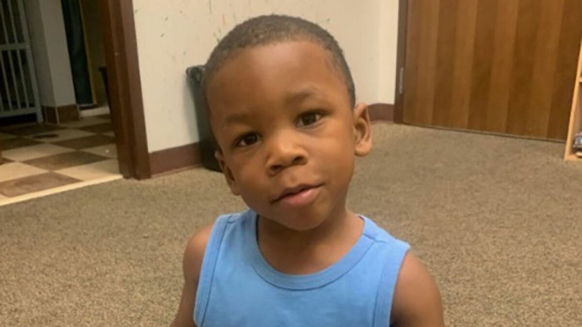 3-year-old boy in temporary foster care sitting indoors, wearing a blue sleeveless shirt, looking at the camera.