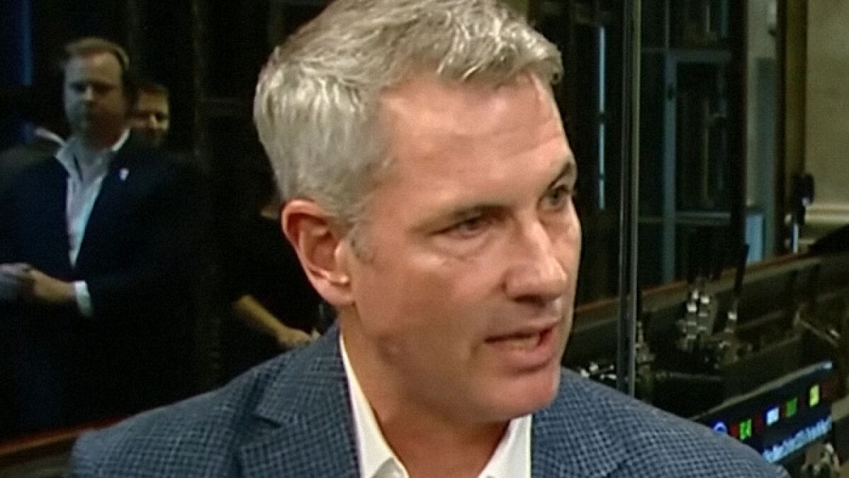 Disgraced CEO Andy Byron speaking during an interview in a suit with a blurred background of people and equipment.