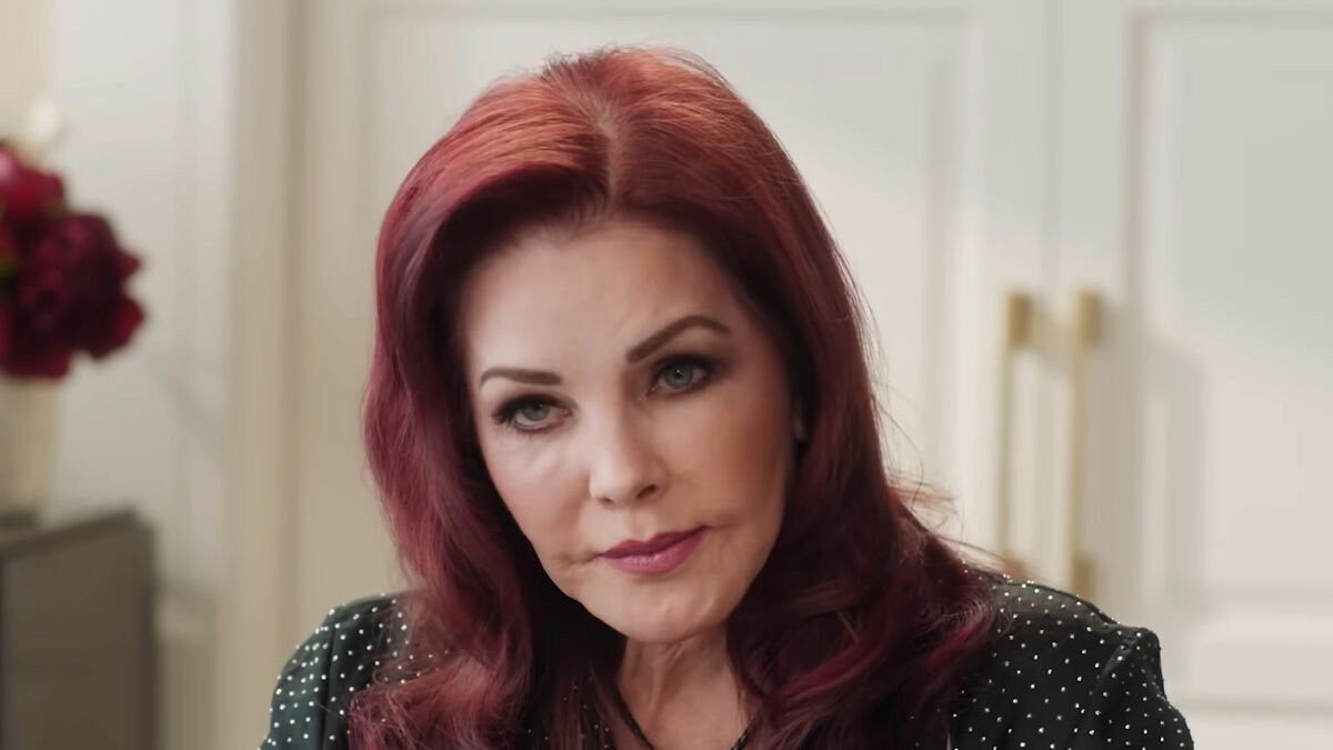 Priscilla Presley, 80, with red hair and a somber expression, seated indoors in a softly lit room.