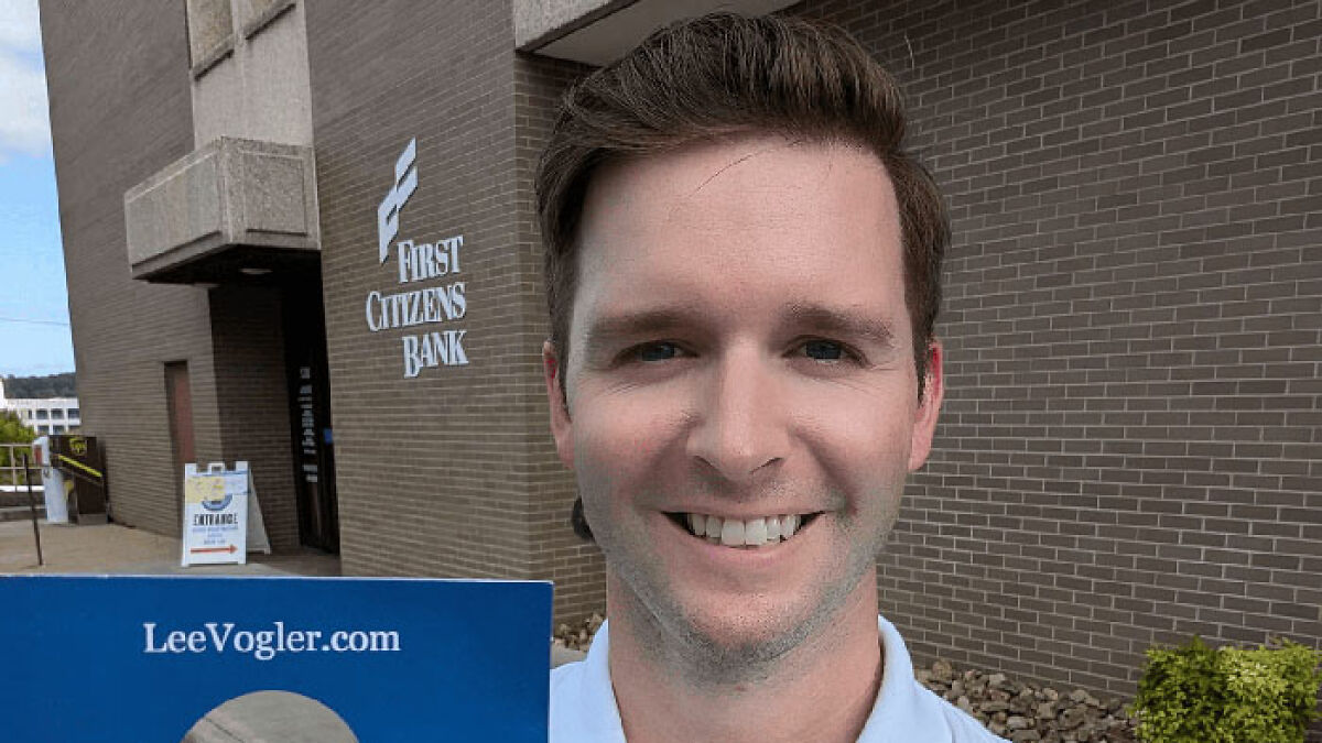 Virginia councilman smiling outdoors holding a campaign sign near a brick building with bank signage visible.