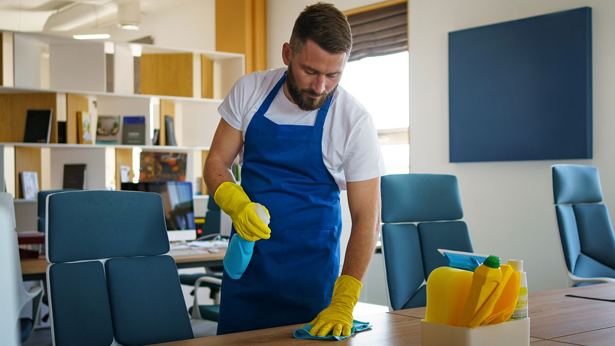 Custodian wearing gloves and apron cleaning office desk, related to boss confronts custodian and feminine hygiene waste bins.
