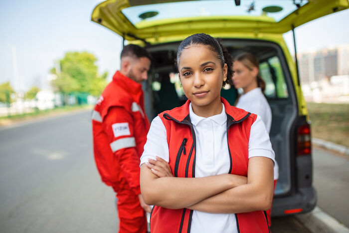 Female first responder standing confidently with arms crossed near ambulance, with two colleagues in the background outdoors.