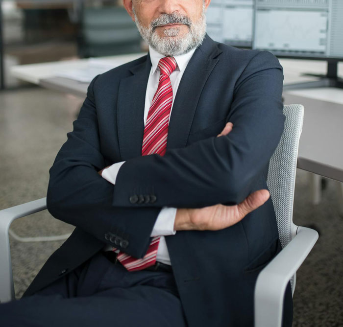 Older man in suit with gray beard sitting with arms crossed in office, portraying a creepy professor mood.