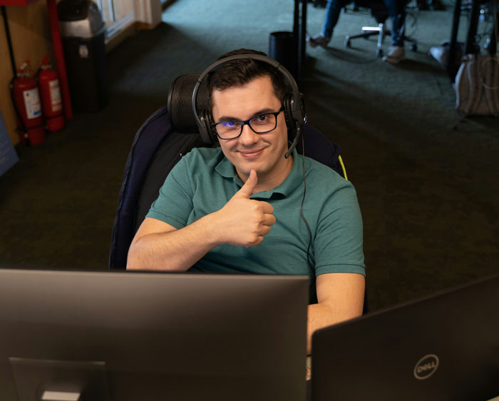 IT professional wearing headset and glasses, sitting at desk with multiple monitors, giving thumbs up in office setting