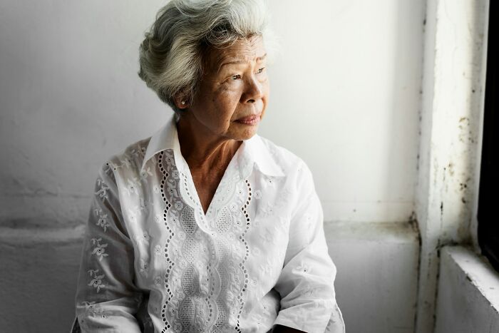Elderly woman in a white blouse looking thoughtfully out a window, reflecting on arrogant people saying leave comments.