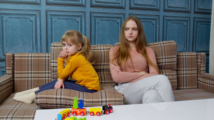 Two girls sitting apart on a couch, showing tension and refusal to accept stepmom in a family setting. Two girls sitting apart on a couch, showing tension and refusal to accept stepmom in a family setting.