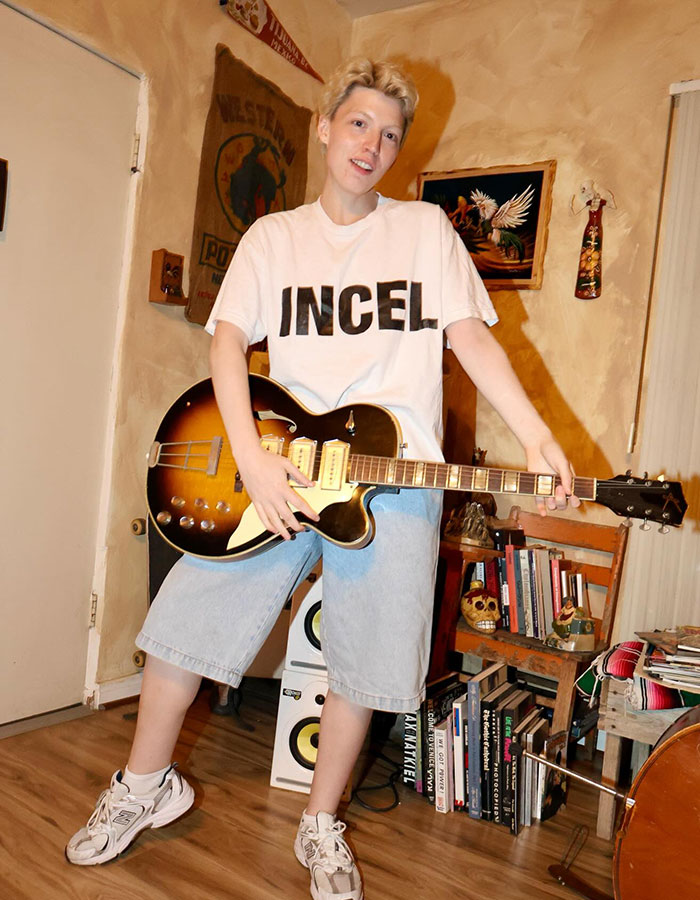 Person wearing a white shirt and shorts playing guitar indoors, representing model pushed to transition and now detransitions. Person wearing a white shirt and shorts playing guitar indoors, representing model pushed to transition and now detransitions.
