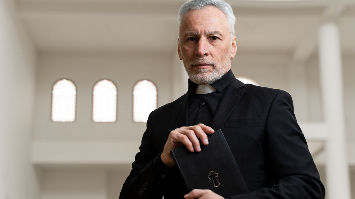 Older male pastor in black clerical attire holding a Bible inside a church during daughter church camp Houston event