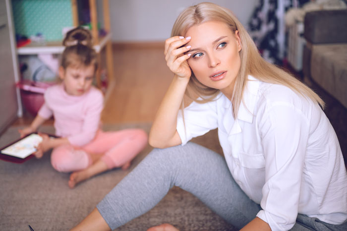Stressed mom sits on floor while child plays in background, reflecting struggles with custody weeks and daycare CPS calls. Stressed mom sits on floor while child plays in background, reflecting struggles with custody weeks and daycare CPS calls.