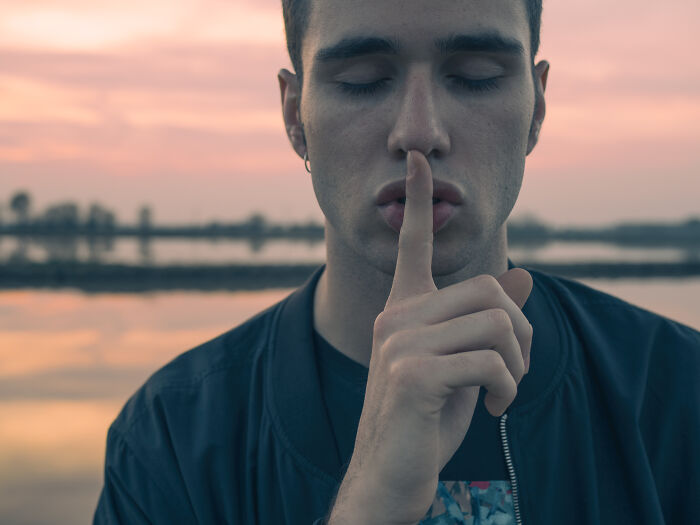 Young man with eyes closed holding finger to lips by calm lake at sunset, representing silent things deaf people experience.