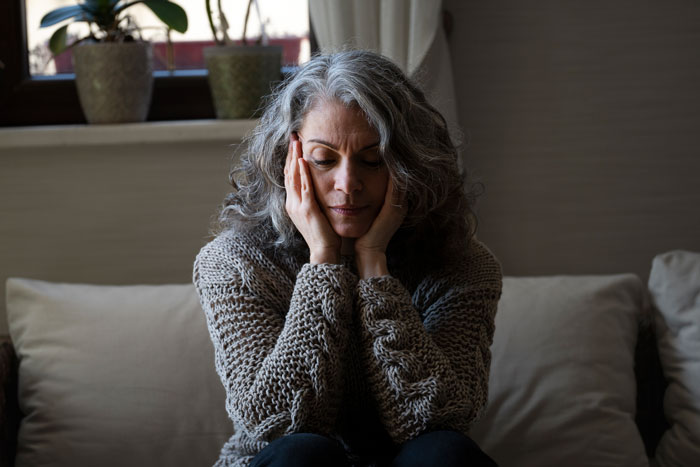 Woman with gray hair sitting on a couch, looking thoughtful and holding her face, representing delivery room baby DNA concerns.