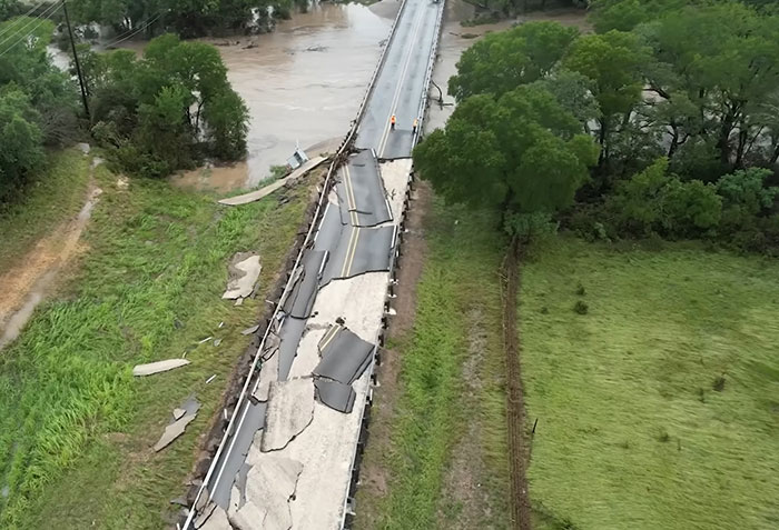 Damaged bridge over floodwaters with two workers inspecting the site, highlighting impact of young flood victim's final message. Damaged bridge over floodwaters with two workers inspecting the site, highlighting impact of young flood victim's final message.
