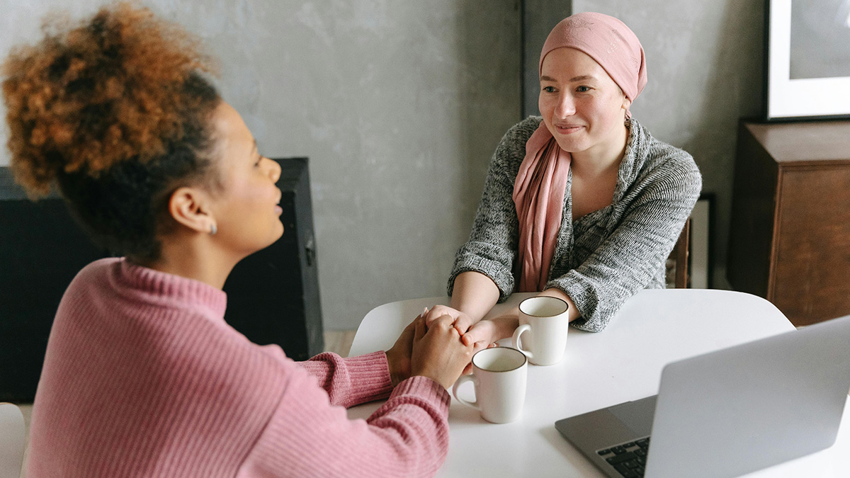 Two women holding hands and sharing a moment with coffee mugs and a laptop on the table showing people helping others.