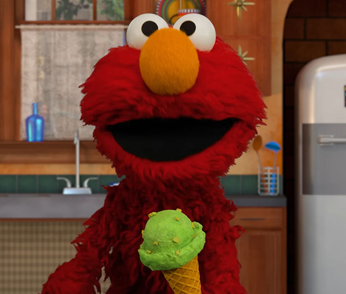 Elmo holding a green ice cream cone in a kitchen setting with a blurred refrigerator and utensils in the background. Elmo holding a green ice cream cone in a kitchen setting with a blurred refrigerator and utensils in the background.