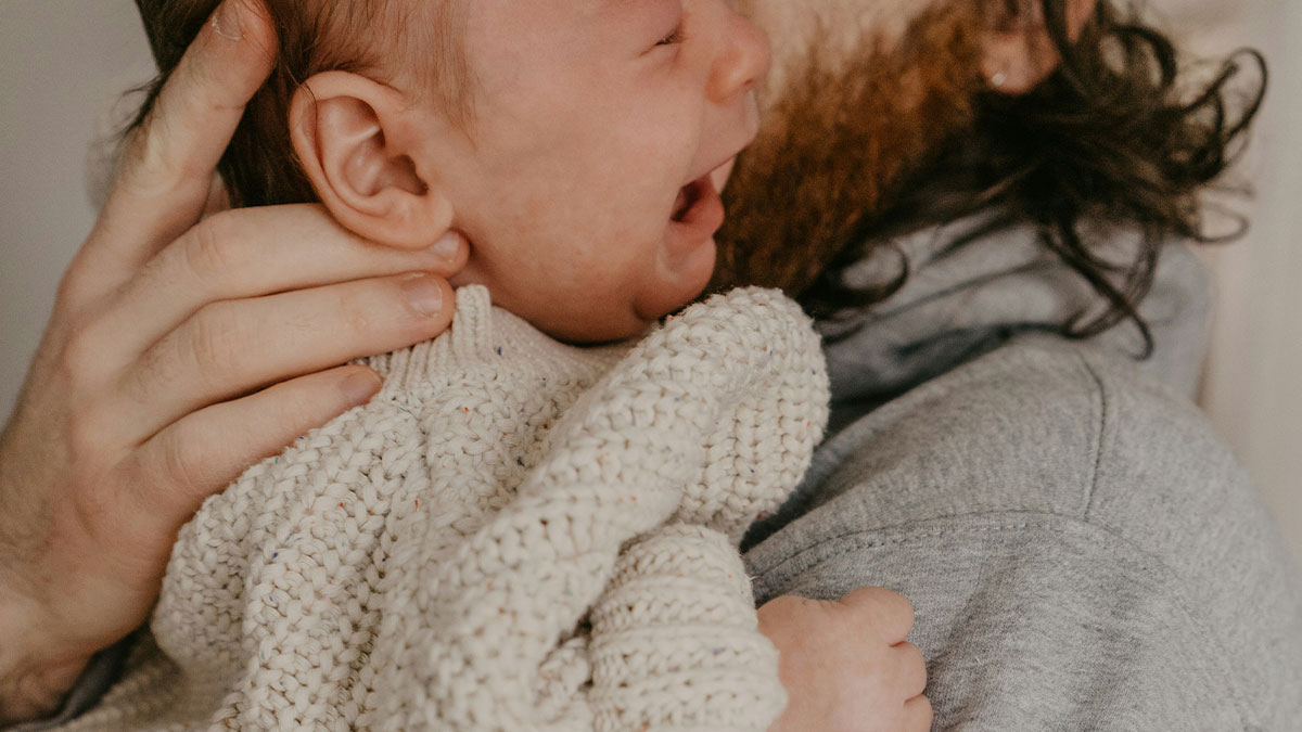 Close-up of a baby wearing a knitted sweater being held by an adult with long hair and a beard.