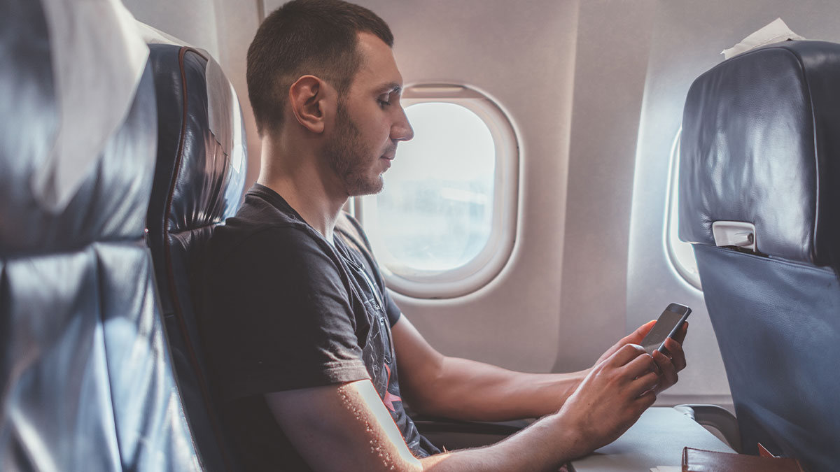 Young man, cancer survivor, sitting in airplane seat by window using smartphone during flight.