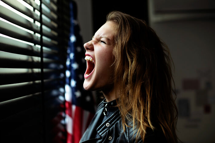 Young woman with long hair shouting by window blinds next to an American flag, relating to escape room employees stories.