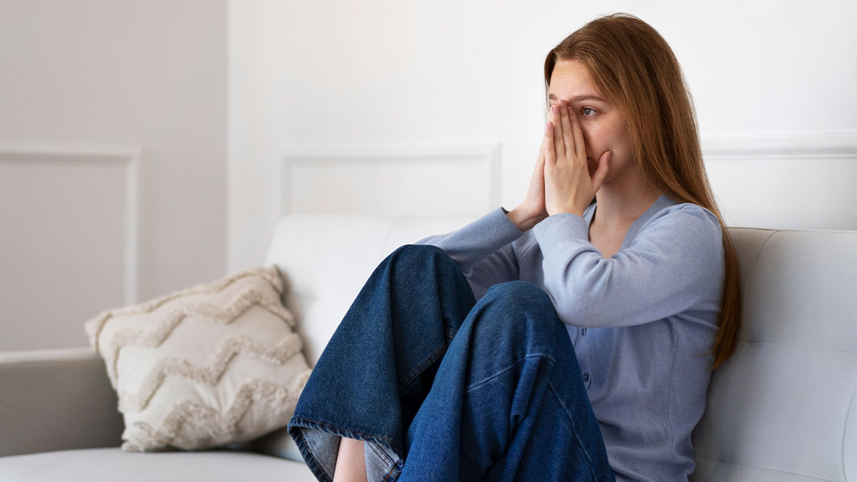 Young woman sitting on couch, revisiting past breakup, covering face with hands, appearing shocked and emotional.