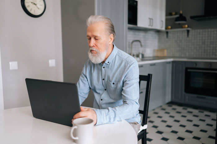 Elderly man with beard using laptop at home, reflecting on false accusation involving wife and father issues.