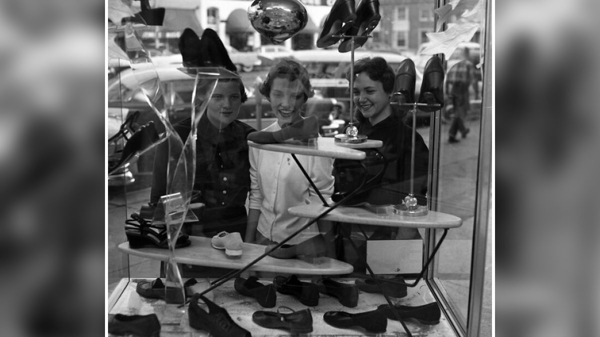 Three women smiling and looking at shoes in a shop window showcasing daily life across the globe in the 50s.