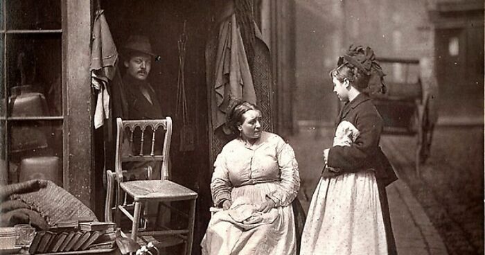 Victorian-era London street scene showing poor residents in worn clothing outside a dilapidated building, illustrating poverty life.