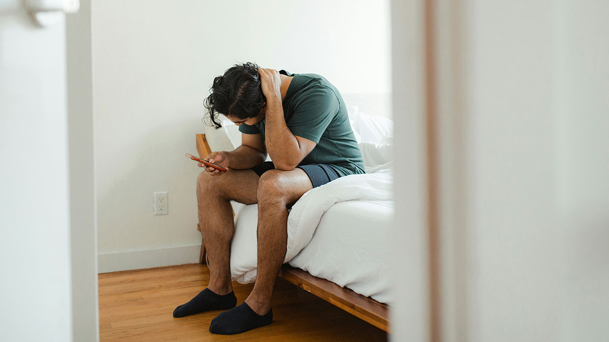 Man sitting on bed looking distressed while holding phone, reflecting pain of fiancee leaving after cancer diagnosis partner.