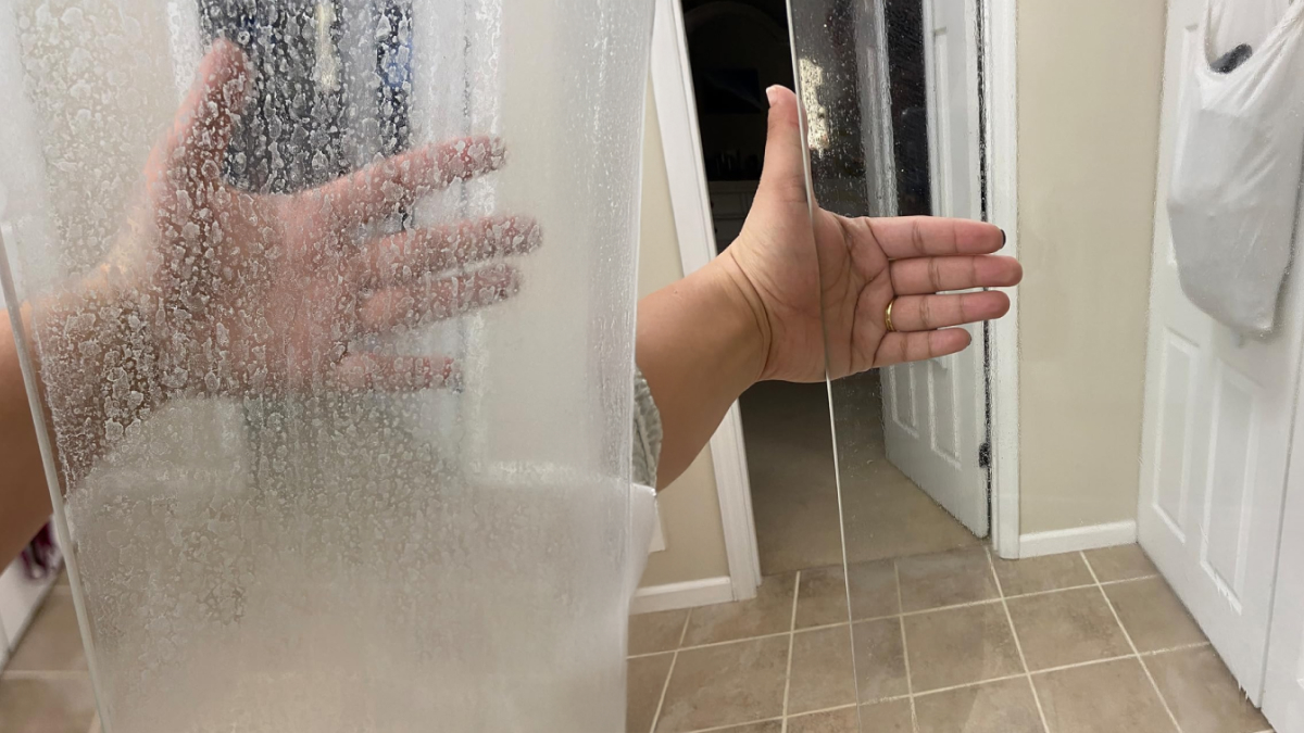 Hand showing comparison of dirty and spotless grout line on transparent cleaning panel in a bathroom.