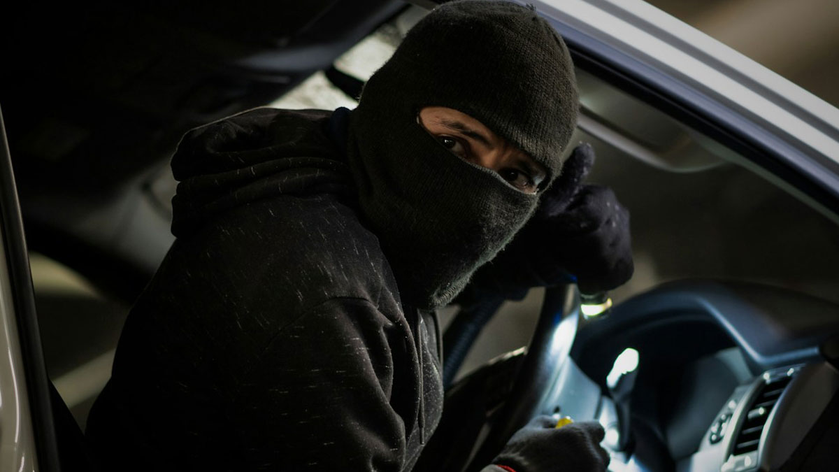 A former home burglar wearing a black mask and gloves inside a car, looking cautiously while attempting a break-in.