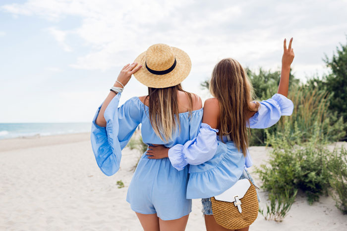 Two women on a beach vacation, one wearing a straw hat, highlighting a friendship trip causing family rift. Two women on a beach vacation, one wearing a straw hat, highlighting a friendship trip causing family rift.