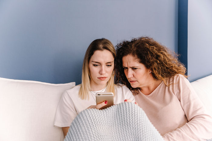 Two women sitting on a couch looking concerned at a smartphone, illustrating vacation rift over friend versus family trip choice. Two women sitting on a couch looking concerned at a smartphone, illustrating vacation rift over friend versus family trip choice.