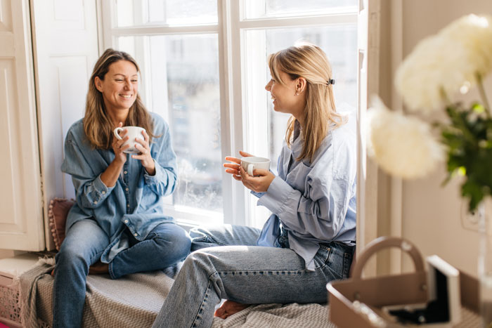 Two women enjoying coffee and conversation by a sunny window, reflecting on single mom and babysitting challenges. Two women enjoying coffee and conversation by a sunny window, reflecting on single mom and babysitting challenges.