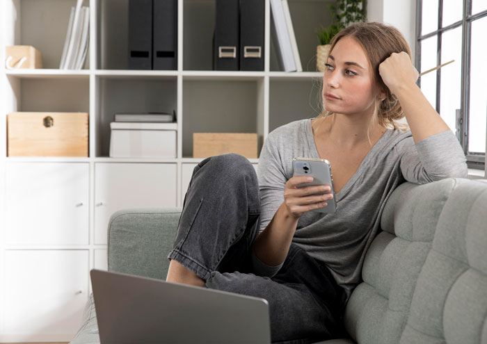 Woman sitting on a couch holding phone looking thoughtful, illustrating single mom facing babysitting challenges. Woman sitting on a couch holding phone looking thoughtful, illustrating single mom facing babysitting challenges.