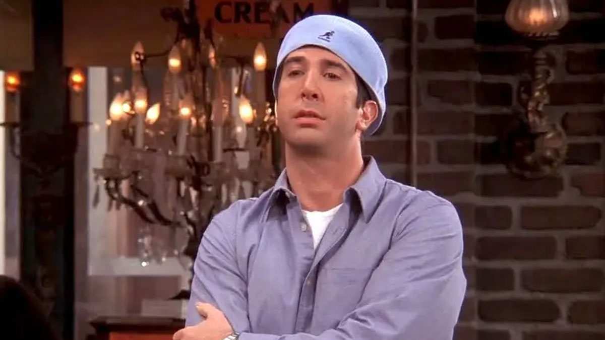 Man in a blue shirt and backward cap inside a cozy room with chandeliers and brick walls, meme from Tamagotchis and VHS era.