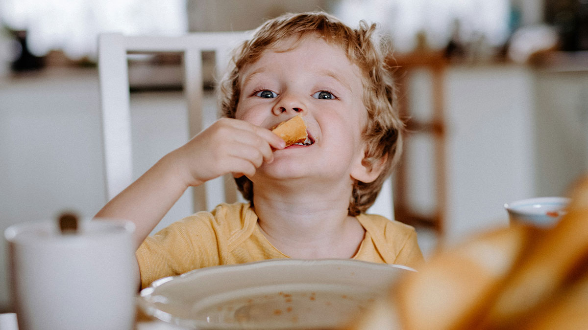Young child enjoying snack time, eating a piece of bread with a joyful expression at the dining table.