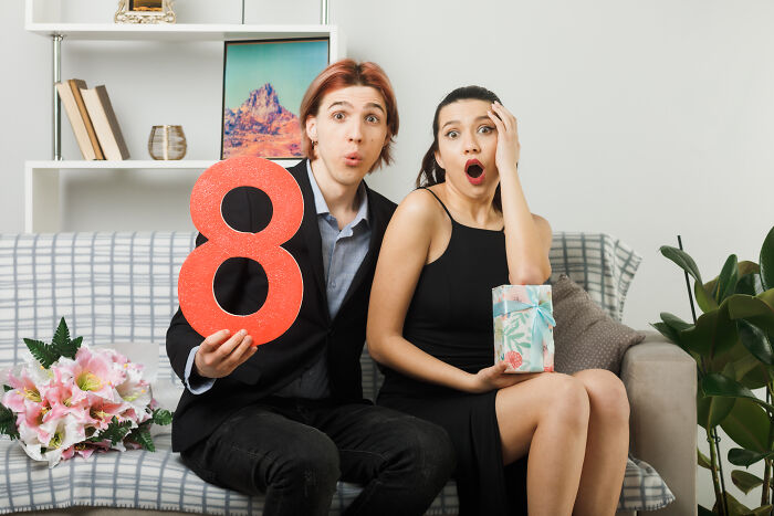Two surprised young adults sitting on a couch showing a red number eight and a gift, illustrating Gen Z compliments.
