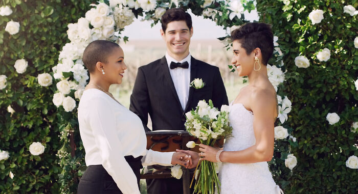 Couple exchanging vows at a wedding ceremony with an officiant, surrounded by white flowers and greenery.