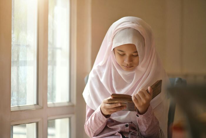 Young girl wearing a white hijab reading a wooden book by a window, illustrating religious myths about Jesus and the Bible.