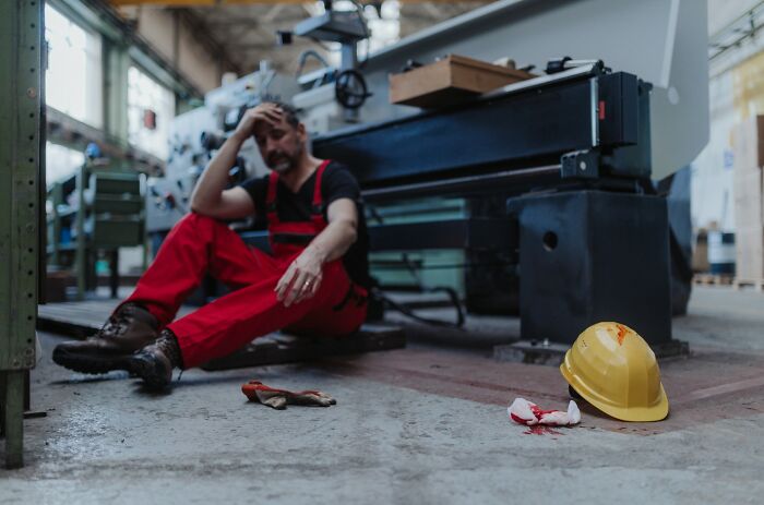 Worker in red overalls sitting on factory floor with hard hat and bloodied bandage nearby, showing regret after arrogant words.