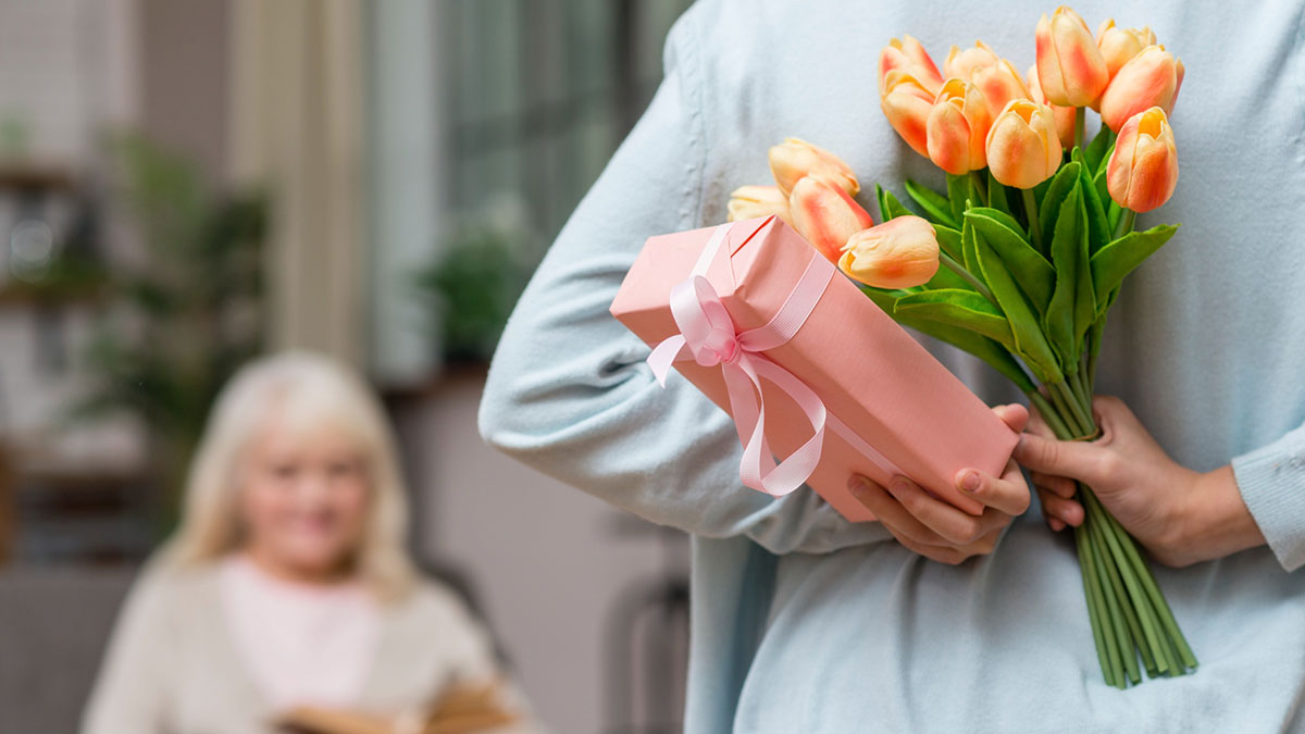 Person holding flowers and a gift behind their back for a Mother's Day surprise with a man expecting a gift from his girlfriend.