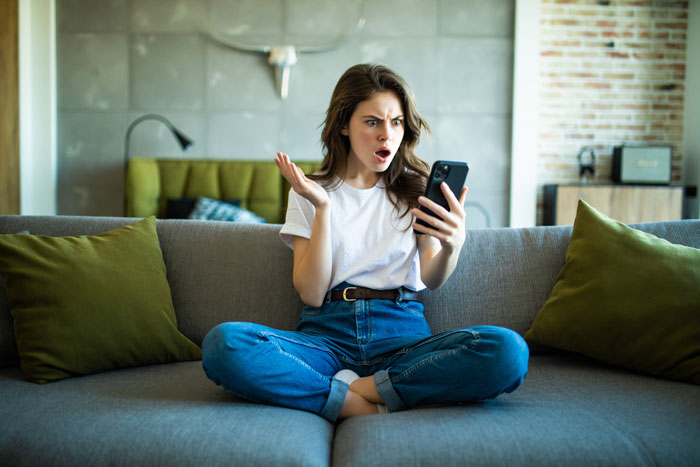 Young woman sitting on couch looking shocked at her phone during gold-digger holiday friend drama in modern living room. Young woman sitting on couch looking shocked at her phone during gold-digger holiday friend drama in modern living room.