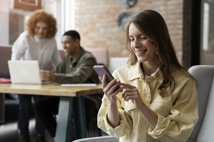 Young woman smiling at phone while two colleagues work together in the background, highlighting oddly specific green flags.