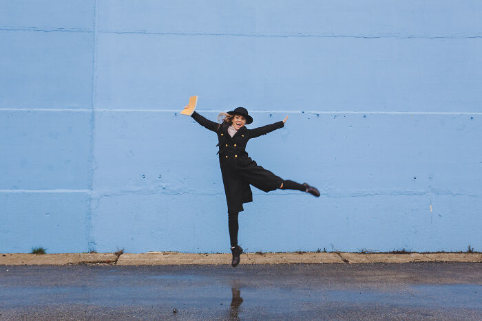 Woman in black coat and hat joyfully jumping near blue wall, illustrating oddly specific green flags in relationships.